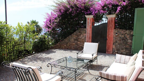 Une terrasse extérieure avec un sol en pierre, une table à plateau en verre, deux chaises blanches rembourrées et un canapé à rayures.