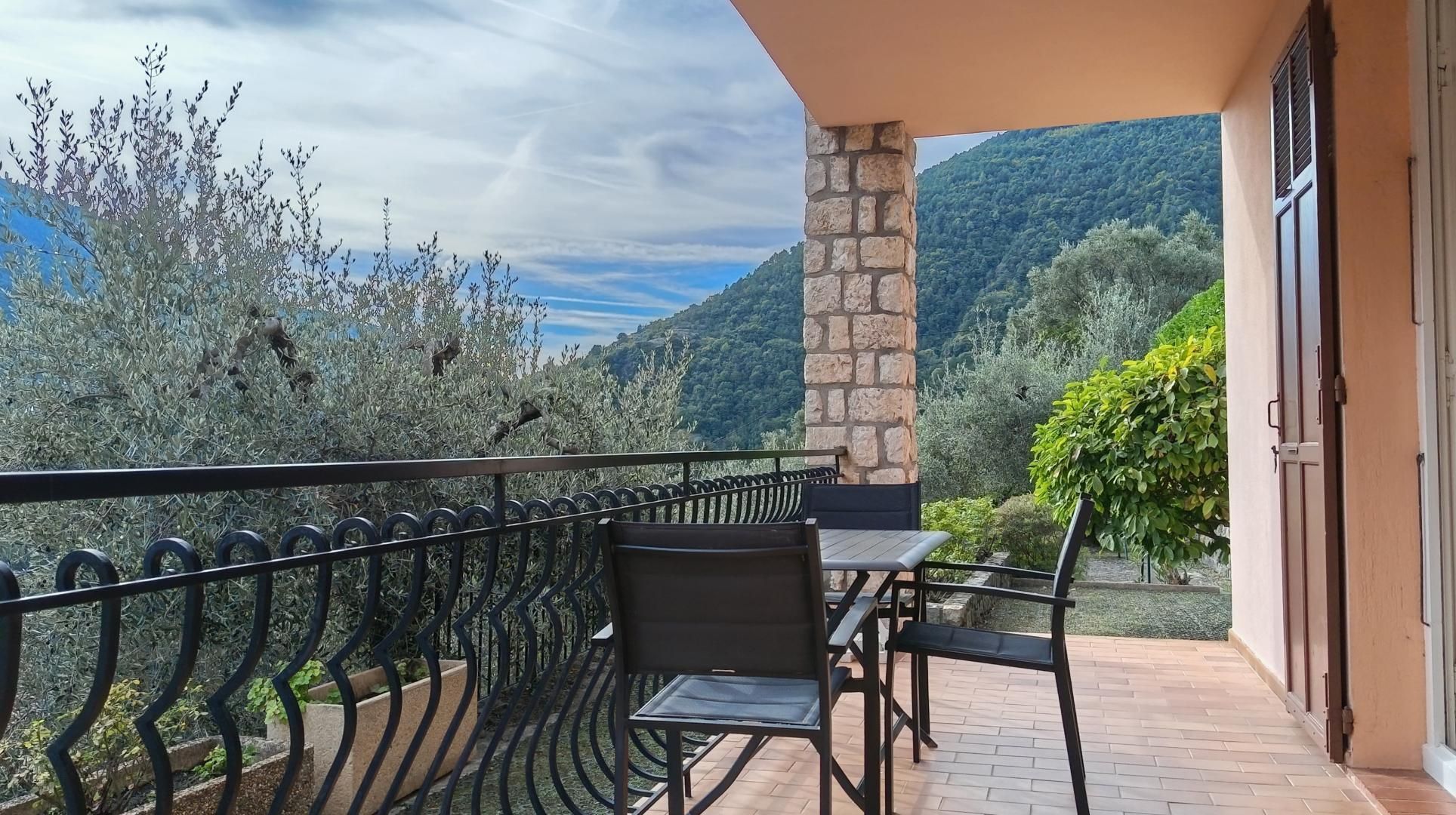 A balcony with a stone pillar, a table, and chairs, overlooking a mountainous landscape.
