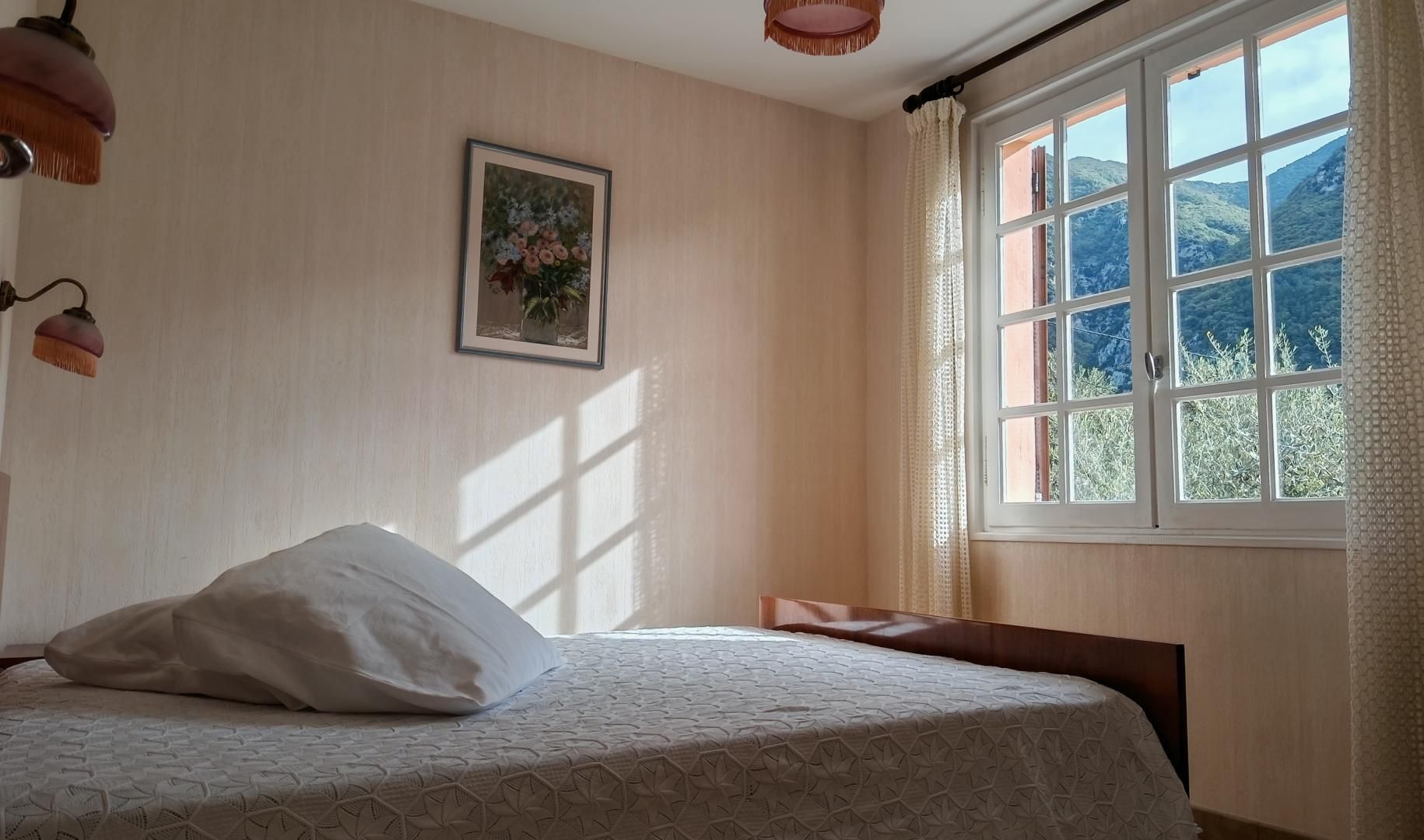 Un dormitorio con cama, ventana con vistas a montañas, un cuadro enmarcado en la pared y una lámpara colgante.