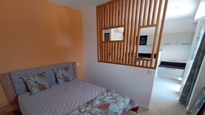 Een slaapkamer met een bed, een houten hoofdbord, een spiegel aan de muur en een kitchenette op de achtergrond.