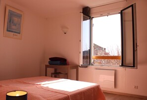 A bedroom with a bed, a bedside table with folded clothes, a lit candle, and a window with an open shutter. - L'Escaou Très - COURSEGOULES (COURSEGOULES)