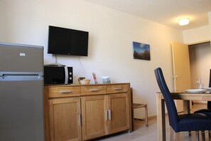 A compact kitchen with a refrigerator, microwave, and a flat-screen TV mounted on the wall. There is a wooden cabinet with drawers and a small table with chairs. A painting hangs on the wall above the table. - L'Escaou Très - COURSEGOULES (COURSEGOULES)