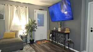 A living room with a grey couch, a flat-screen TV mounted on the wall, a small side table with a lamp and books, and a potted plant.