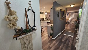 A hallway leading to a kitchen with a dark wooden floor, a white wall with a macrame decoration, and a mirror.
