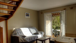 A cozy living room with a sofa, a small table, and a dining table with chairs. A wooden staircase is visible on the left.