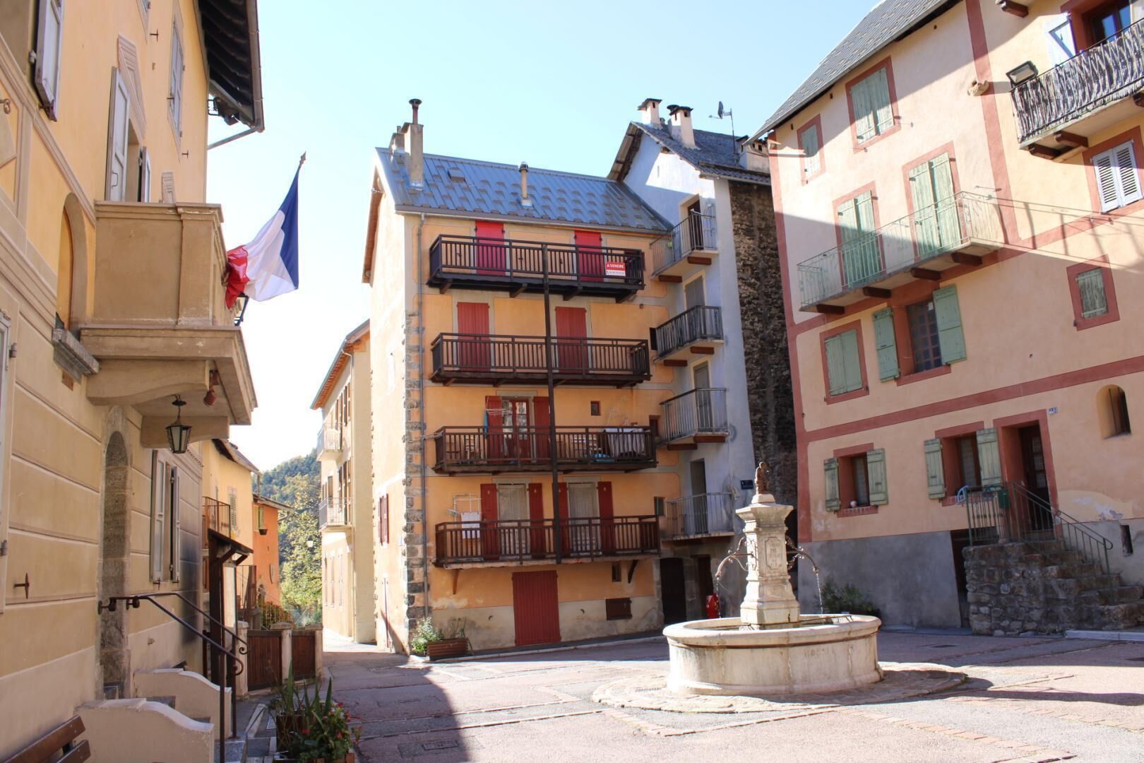 Une rue étroite, bordée de bâtiments des deux côtés, avec une fontaine au centre et un drapeau sur le côté gauche.