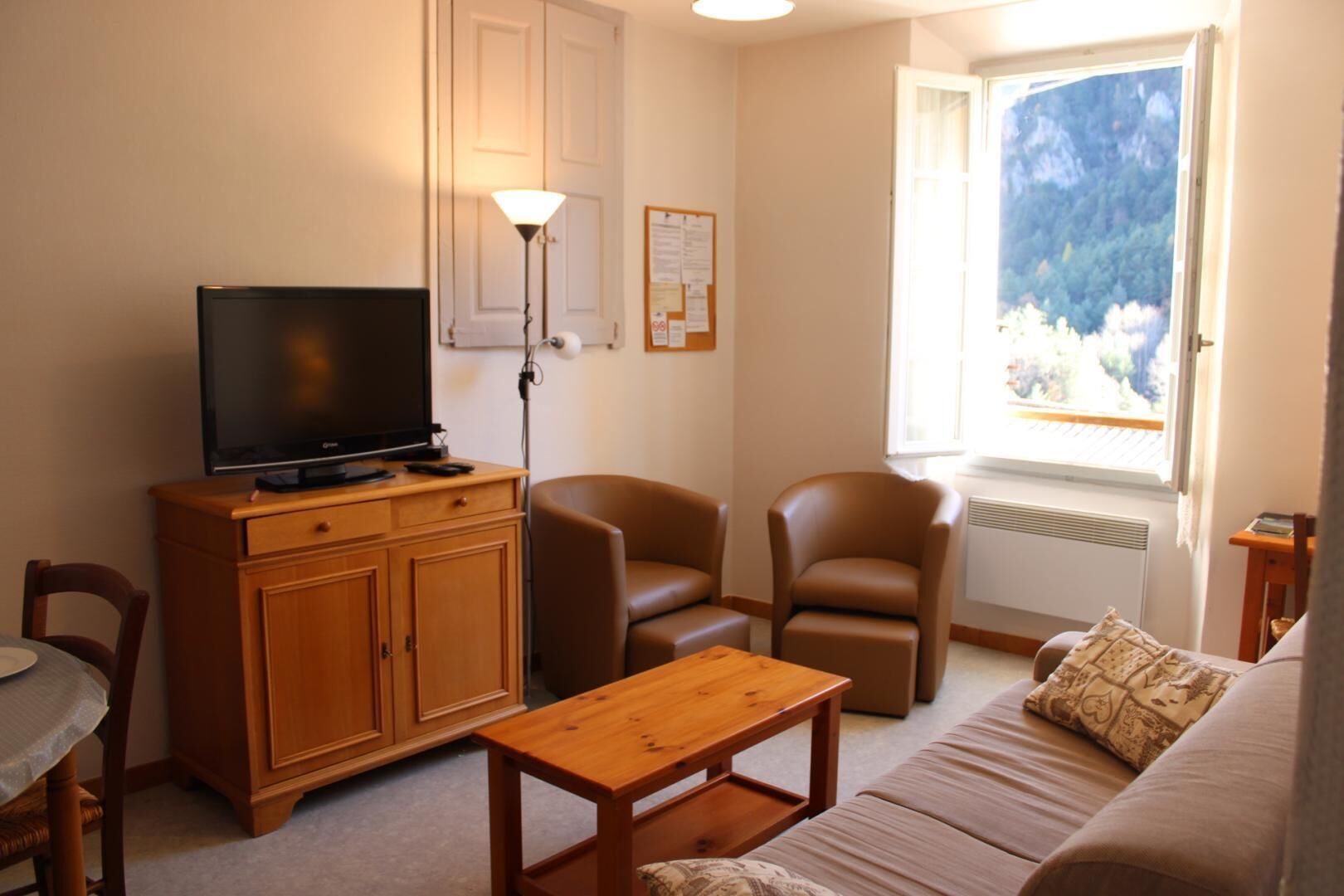 A living room with a television on a wooden cabinet, two leather chairs, a wooden coffee table, a sofa with a patterned cushion, and a window with a mountain view.