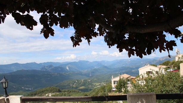 A scenic view from a balcony with a wooden railing, overlooking a valley with mountains in the distance and a building with a tiled roof.