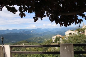 A scenic view from a balcony with a wooden railing, overlooking a valley with mountains in the distance and a building with a tiled roof.