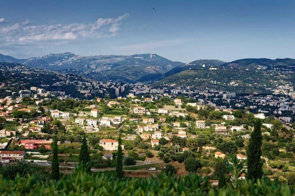 Une vue panoramique d’une ville dont les bâtiments sont dispersés sur un paysage vallonné, entourée de verdure et avec des montagnes en arrière-plan.