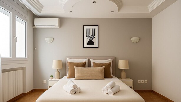 A hotel room with a large bed, two bedside lamps, a wall-mounted air conditioner, and a framed artwork above the bed. - Spacieux Appartement à Hydra (Timokten)