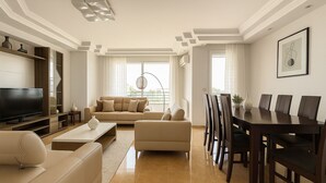 A modern living room with a flat-screen TV, beige sofas, a dining table with chairs, and a large window with curtains. - Spacieux Appartement à Hydra (Timokten)