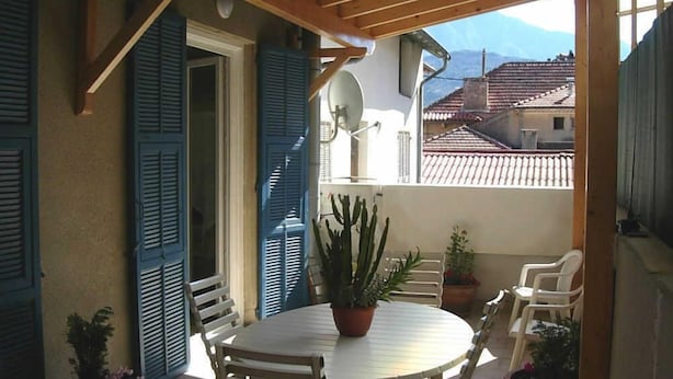 Een overdekt terras met een ronde tafel en stoelen, potplanten en blauwe luiken aan de muren.