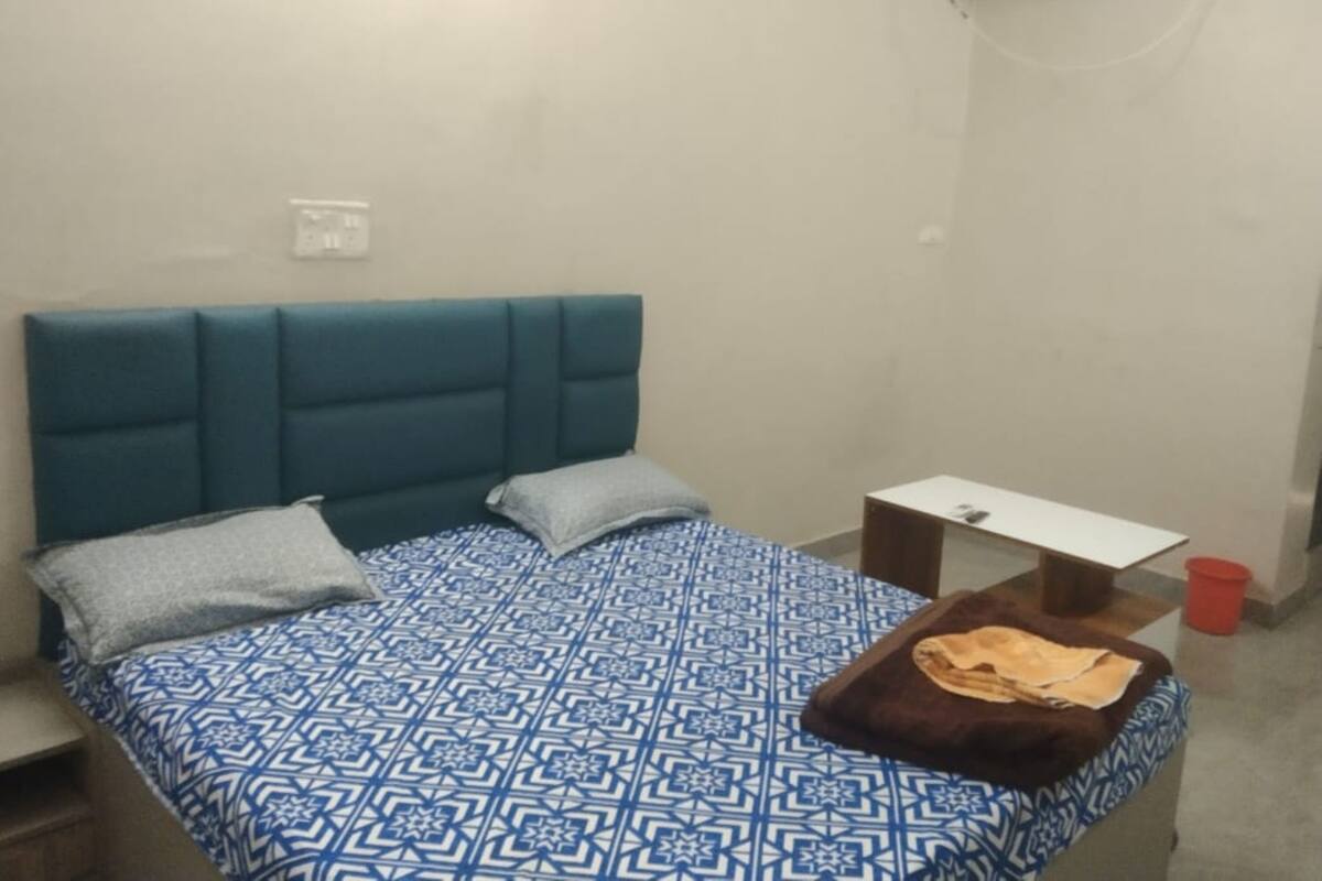Una cama individual con un edredón azul estampado, un cabecero color turquesa y dos almohadas. Hay una mesita de noche de madera con una lámpara, un bote de basura rojo y una mesa blanca con un mantel marrón.