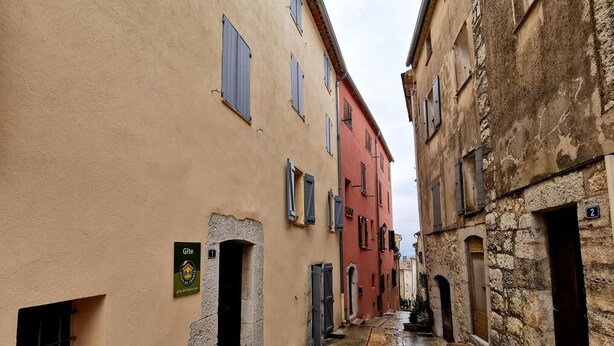 A narrow street with buildings on either side, featuring stone and plaster facades, small windows, and numbered doors.