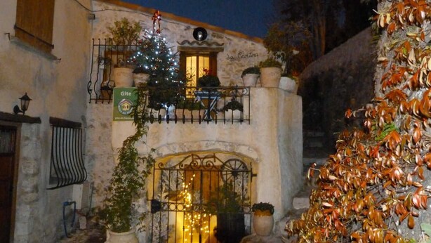 Un bâtiment décoré d’un sapin de Noël et de guirlandes lumineuses, une enseigne indiquant « Gîte », et un mur de pierre orné de plantes.