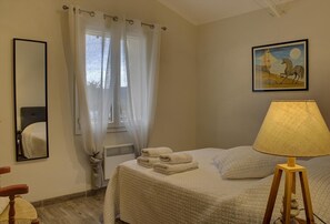 A bedroom with a bed, a bedside lamp, a mirror, a window with curtains, and a painting on the wall.