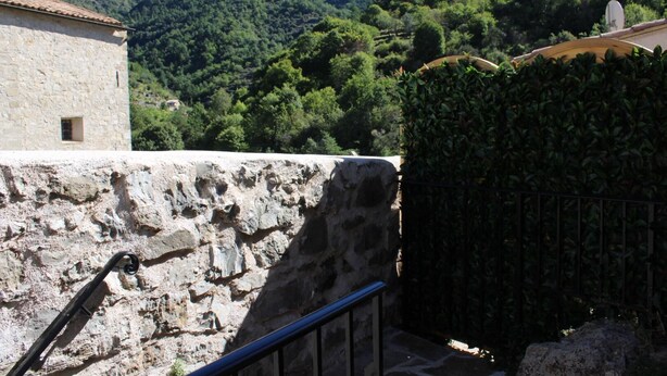 A stone wall with a metal railing, a building with a tiled roof, and a lush green hillside in the background.