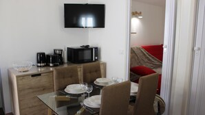 A dining area with a glass table, brown chairs, a TV mounted on the wall, a microwave, and a coffee maker on a wooden cabinet.