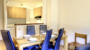A dining area with a glass table, four blue chairs, and a wooden chair. A kitchen with wooden cabinets and a stove is visible in the background.