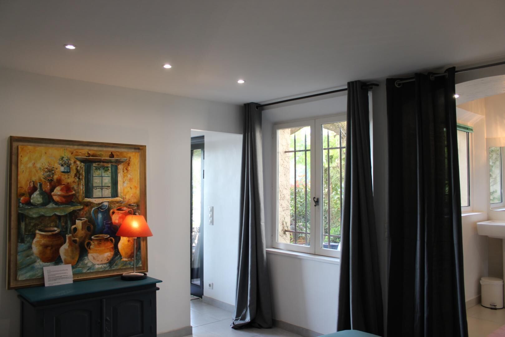 A room with a painting of pottery, a lamp, a window with curtains, and a sink.