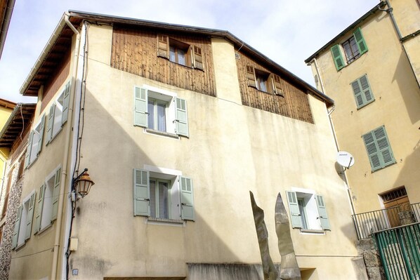 A multi-story building with a beige facade, wooden upper floors, and green shutters on windows.