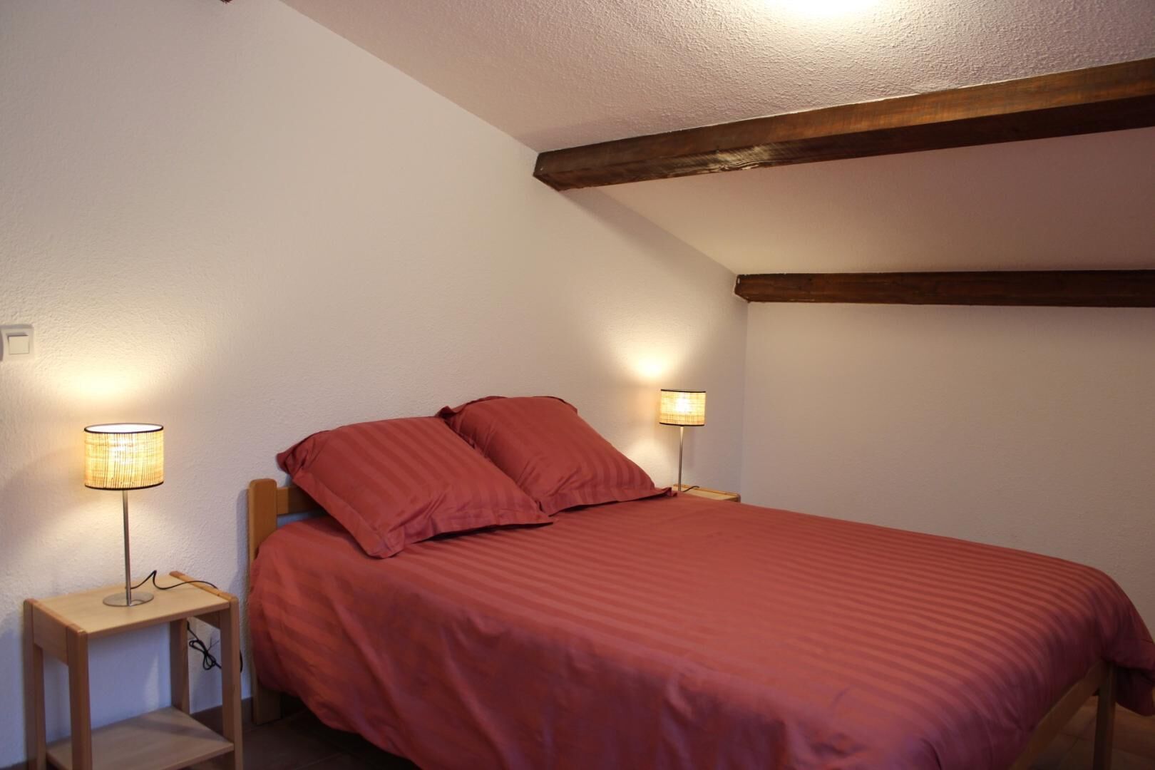 A bedroom with a single bed, two bedside lamps, and a wooden ceiling beam.