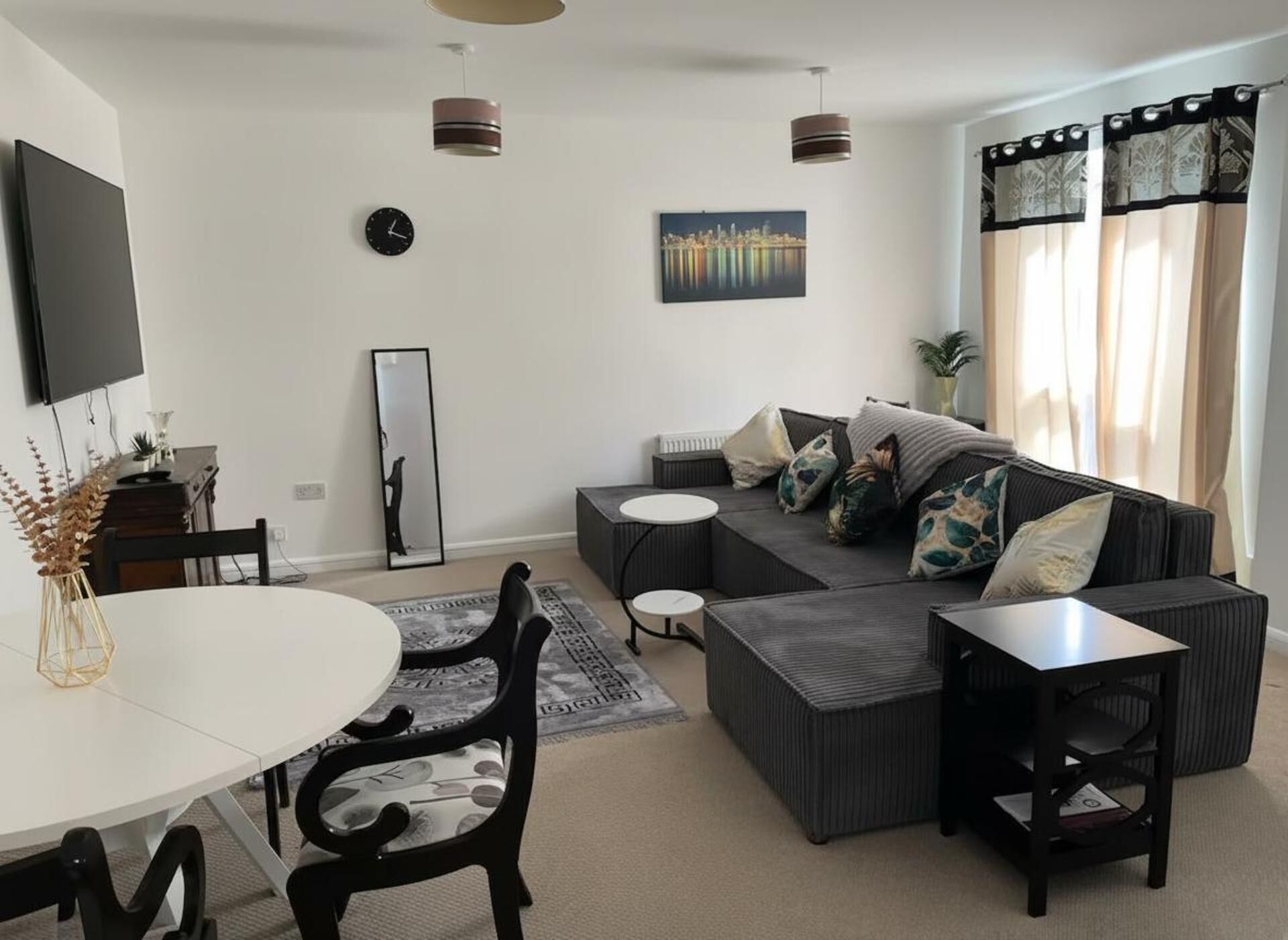A modern living room with a sofa, coffee table, and chairs. There is a flat-screen TV mounted on the wall, a clock, and a painting.