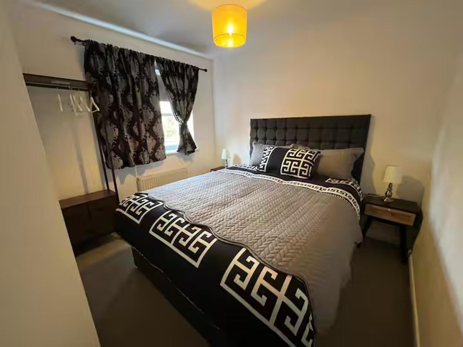 A bedroom with a bed, a nightstand, a lamp, a window with curtains, and a radiator.
