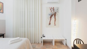 Ett hotellrum med en säng, en vit bänk, en väggmonterad luftkonditioneringsanläggning och en målning som föreställer en dansare.