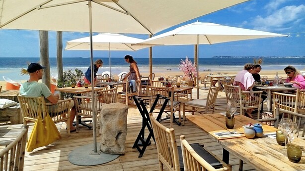 Un área de comedor al aire libre con mesas y sillas de madera, sombrillas grandes y vistas a la playa y el océano.