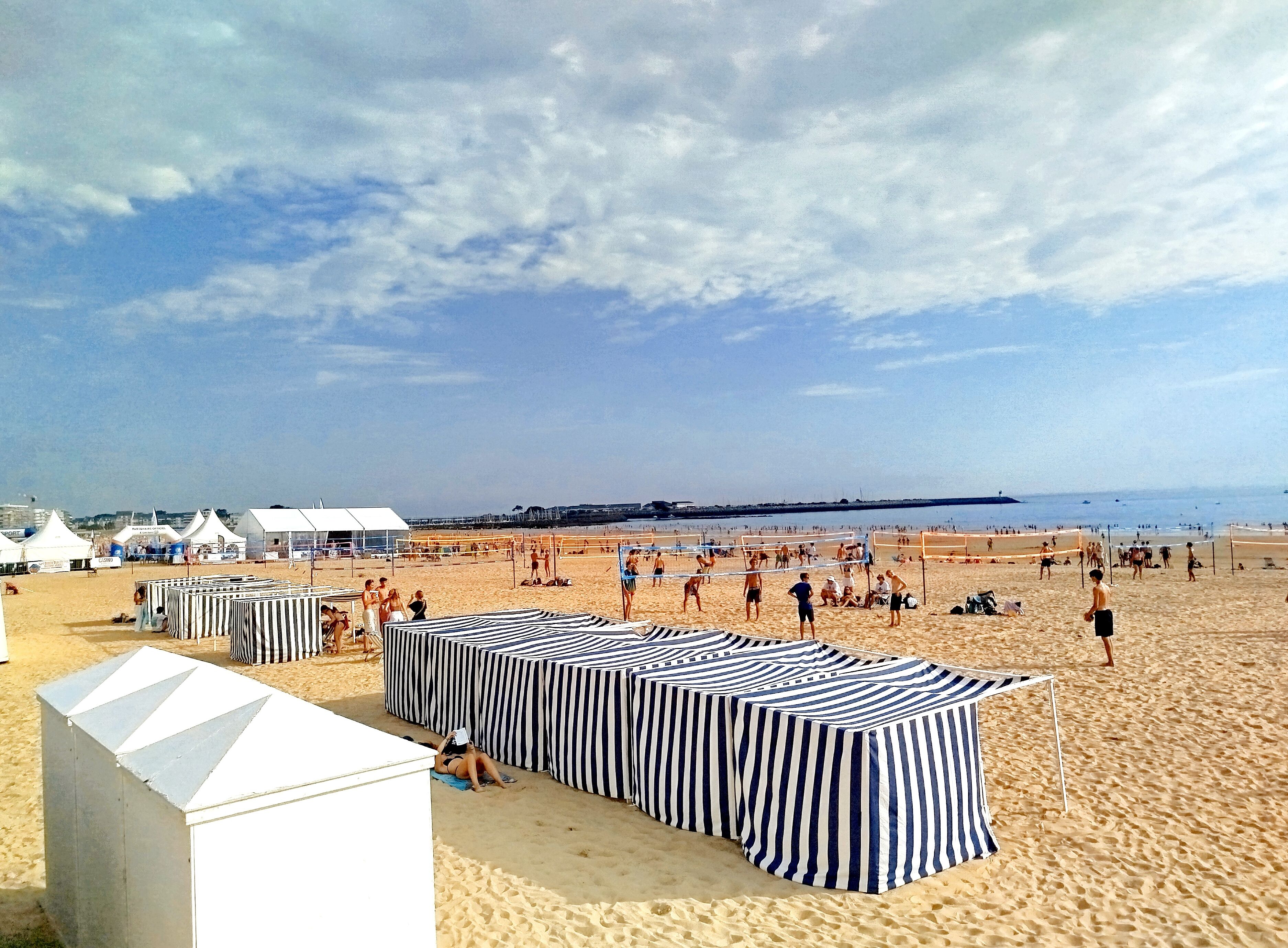 Ein Strand mit gestreiftem Zeltdach und Sonnensegeln, feiner Sand und ein klarer Himmel.