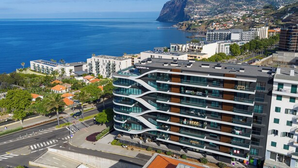 A modern multi-story building with a unique zigzag design, located near a coastal area with a view of the ocean and a mountain in the background. - Oceanview Pearl Apartment (Funchal)