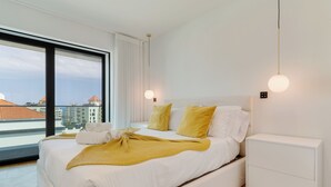 A modern bedroom with a large bed, white bedding, yellow pillows, and a view of buildings through a large window. - Oceanview Pearl Apartment (Funchal)