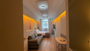 A narrow hotel room with a bed, a sofa, a small table, and a TV. - Monastiraki 303 Superhost Hub (Athens)