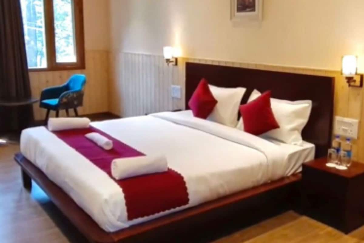 Ein ordentlich bezogenes Bett mit weißer und roter Bettwäsche, ein Holz-Kopfteil und ein Nachttisch mit Lampe und Wasserflaschen.