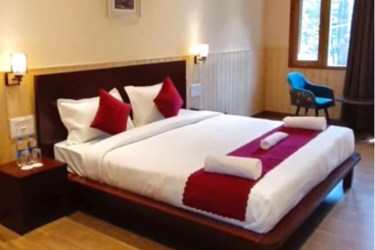 Ein Hotelzimmer mit einem hölzernen Bett, weißer Bettwäsche, roten Kissen und einer roten Tagesdecke. Es gibt einen Sessel und einen Nachttisch mit Lampe und einer Wasserflasche.