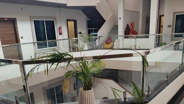 A modern indoor space with a glass railing, a central potted plant on a pink stool, and a staircase in the background. - Agralok Dham (Ujjain)