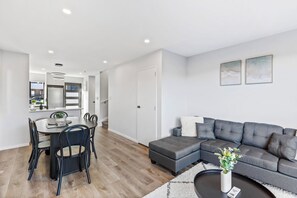A modern living room with a grey sectional sofa, a dining table with chairs, and a kitchen area visible in the background. - Stylish 4BR Townhouse in Suburbs by Zodiak (Auckland)