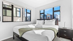 A modern bedroom with a large bed, two windows, a bedside table, and a view of buildings outside.