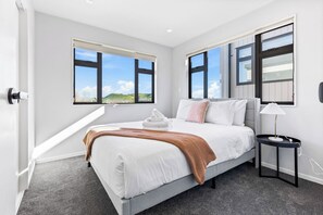 A modern hotel room with a large bed, a bedside table with a lamp, and a view of the outdoors through large windows. - Stylish 4BR Townhouse in Suburbs by Zodiak (Auckland)