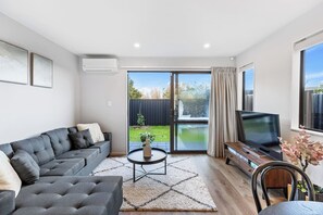 A modern living room with a grey sofa, a coffee table, a flat-screen TV, and a sliding glass door leading to a backyard. - Stylish 4BR Townhouse in Suburbs by Zodiak (Auckland)
