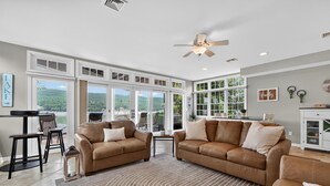 A spacious living room with two brown leather sofas, a patterned rug, and large windows offering a view of greenery.