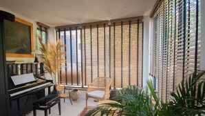 A room with a piano, wicker chairs, a large window with blinds, and potted plants. - Carqueiranne: Charming Villa with Pool, Sea View, for 8 Guests (Carqueiranne)