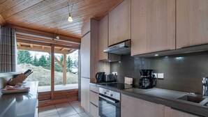 Cocina moderna con armarios de madera, encimera de acero inoxidable, cafetera y vista a un área boscosa a través de un ventanal grande.
