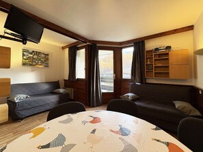 Une pièce chaleureuse avec deux canapés sombres, une petite table recouverte d’une nappe colorée, un téléviseur à écran plat fixé au mur et une bibliothèque.