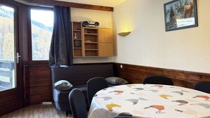 A dining area with a round table covered by a patterned tablecloth, surrounded by chairs. A wooden cabinet with shelves is against the wall, and a painting hangs above the table. - Spacious studio 6 ppl, ski-in/ski-out, Free Wifi (Les Orres)