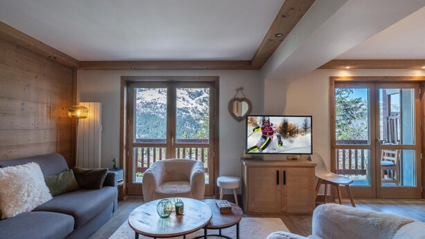 Un salón acogedor con sofá, sillones, una mesa de centro y un televisor que muestra un evento deportivo. Amplias ventanas ofrecen vistas a un paisaje montañoso nevado.