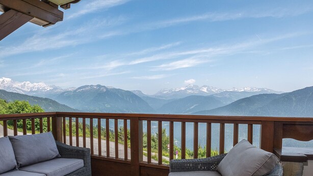 Un balcon agrémenté de meubles en osier et d’une table préparée pour le petit-déjeuner, offrant une vue sur des montagnes enneigées.