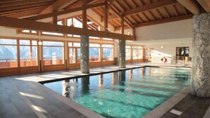 Une piscine intérieure agrémentée de piliers en pierre, de poutres en bois et de grandes fenêtres offrant une vue sur les montagnes.